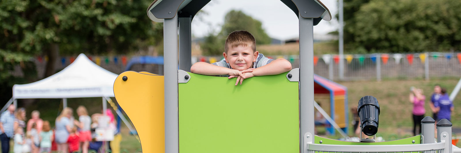 A young boy is standing inside a playhouse on top of a play structure at a playground. He is leaning on his arms on the side and looking and smiling at the camera.